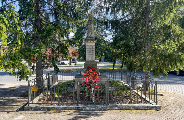 Figura Matki Bożej Pocieszycielki Strapionych za metalowym ogrodzeniem, u stóp czerwone kwiaty