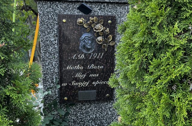 Granitowa kapliczka z 1911 roku w Michałowie-Grabinie z tablicą i kwiatami, między tujami
