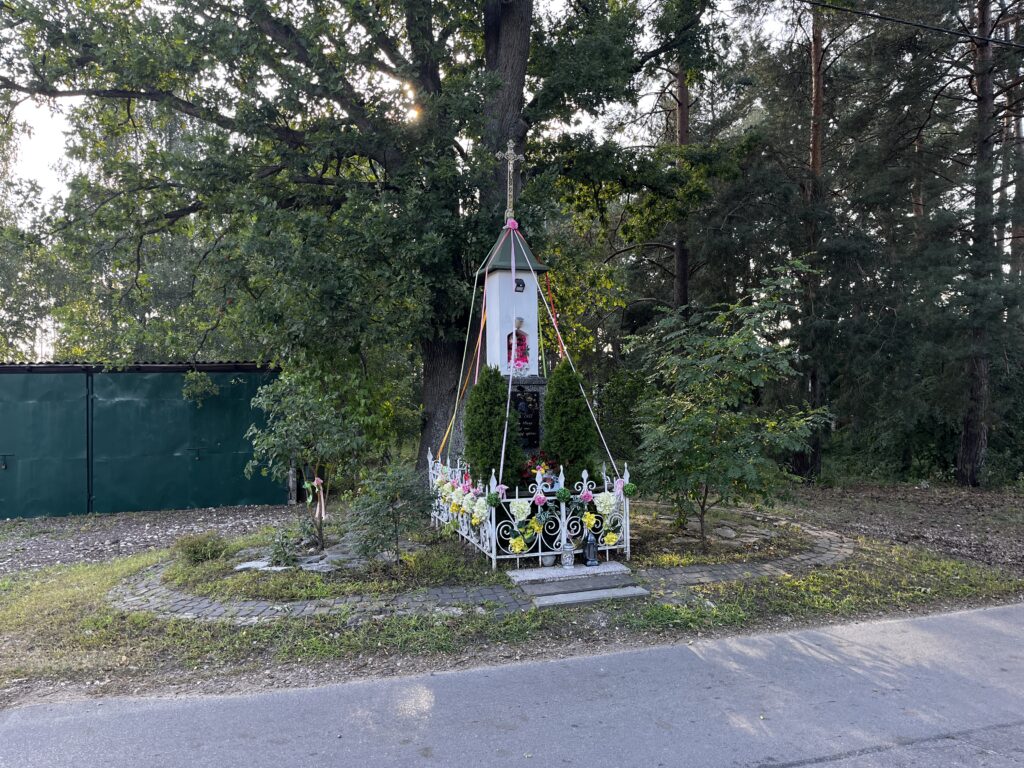 Kapliczka z 1911 roku przy drodze w Michałowie-Grabinie, za białym ogrodzeniem, pod dużym drzewem