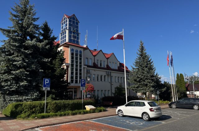 Wejście do Urzędu Gminy Nieporęt z parkingiem, świerkami i masztami z flagami