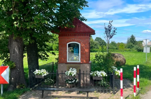 Red-brick wayside shrine from 1900 with a figure of Our Lady, behind a fence with flower boxes, shaded by trees by the road